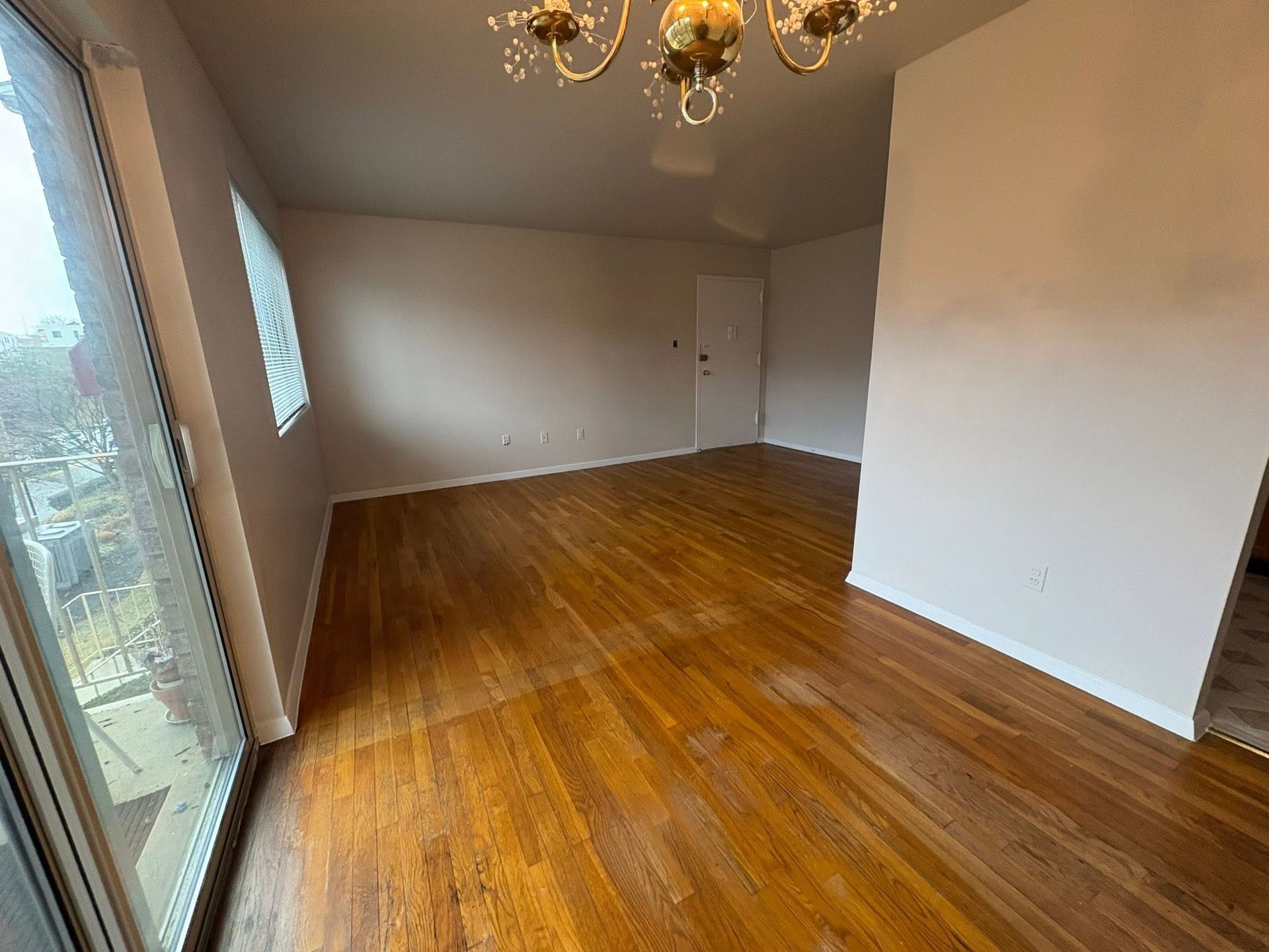 Empty room with hardwood floors, beige walls, and a chandelier near a sliding glass door.