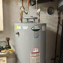 Gray water heater in a utility closet with exposed pipes and a vent pipe on top.