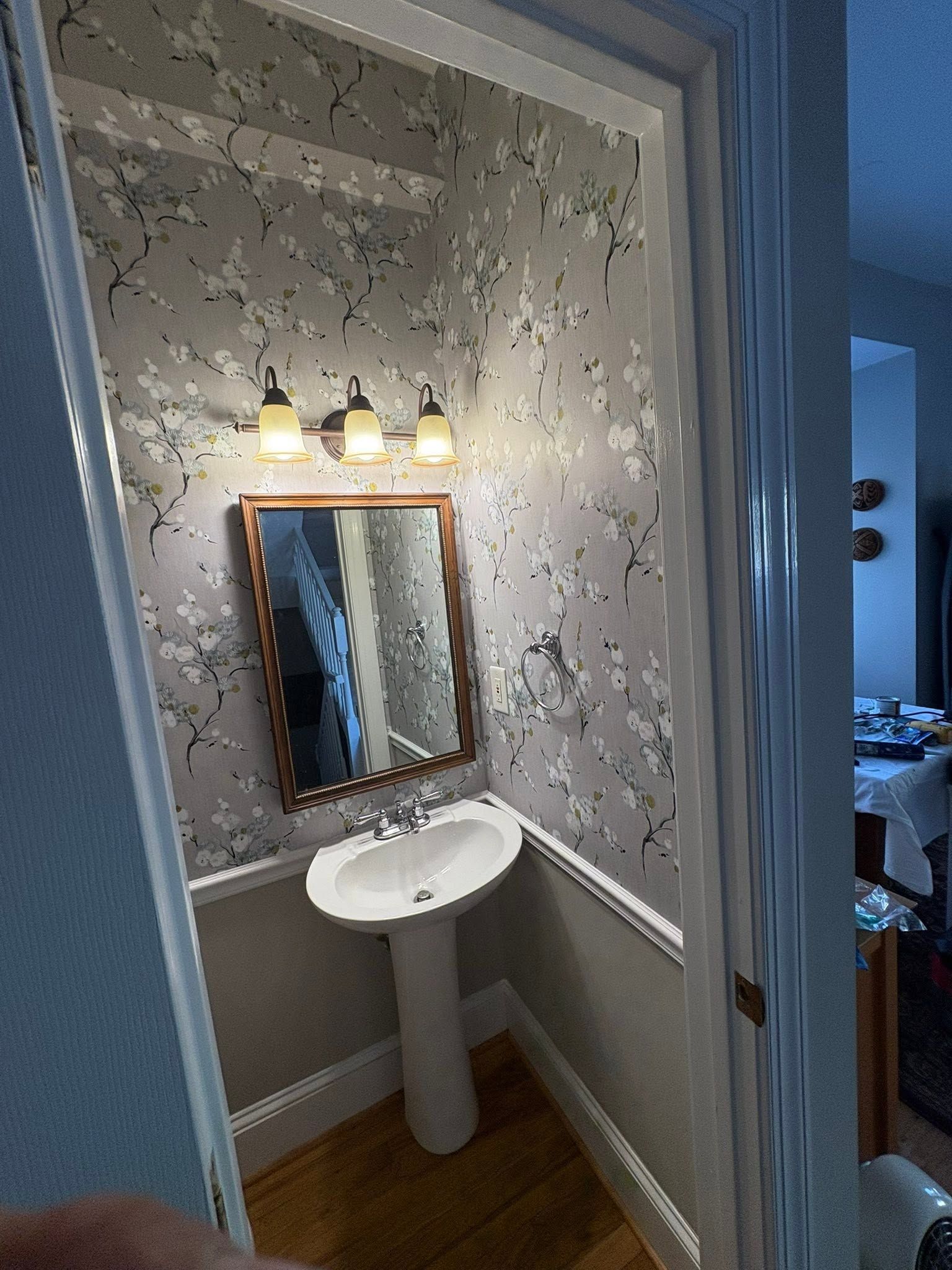 Small bathroom corner with pedestal sink, mirror, wall light, and floral wallpaper