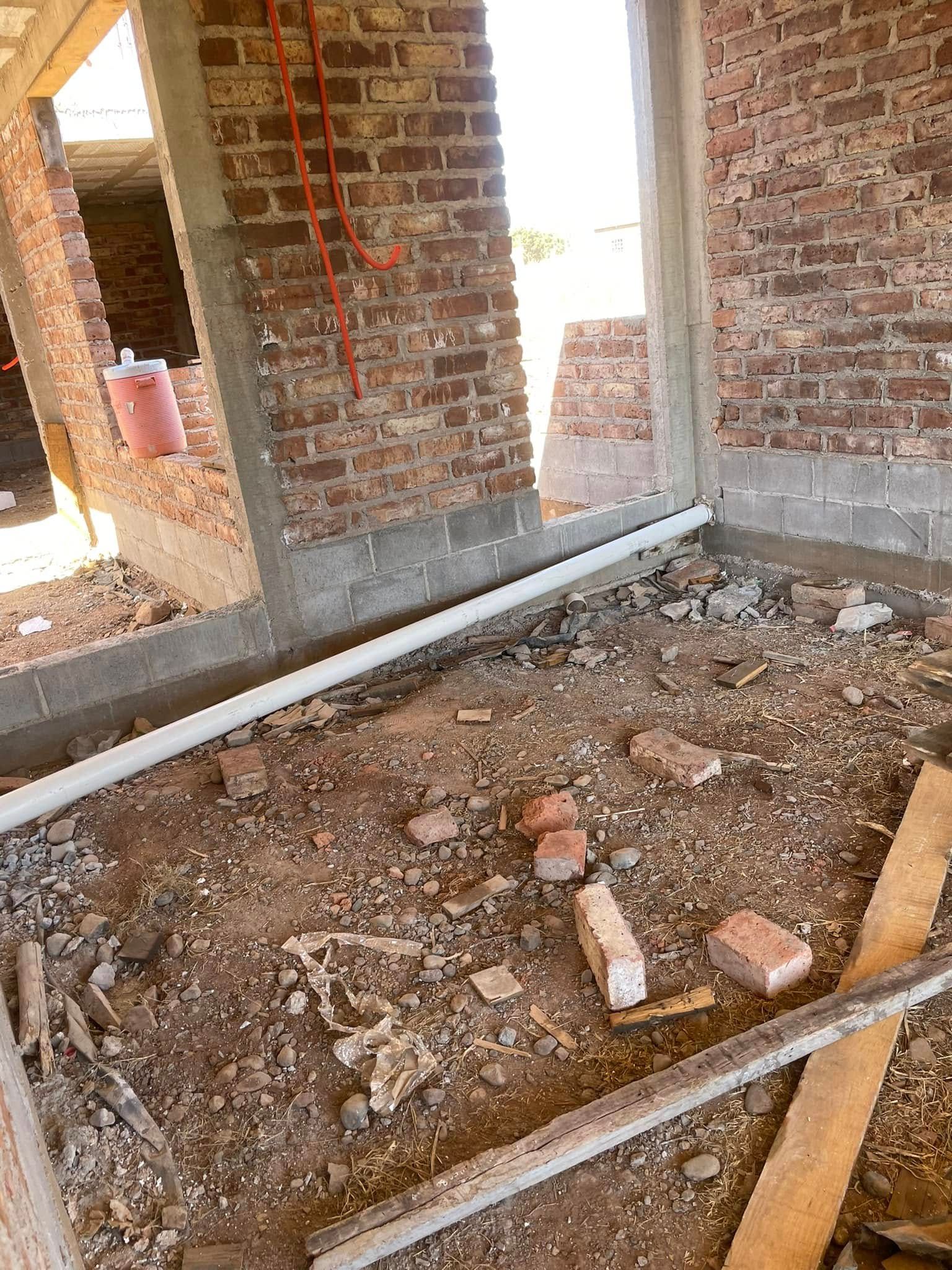 Unfinished brick room with exposed concrete floor, debris, and a wooden plank across the foreground.