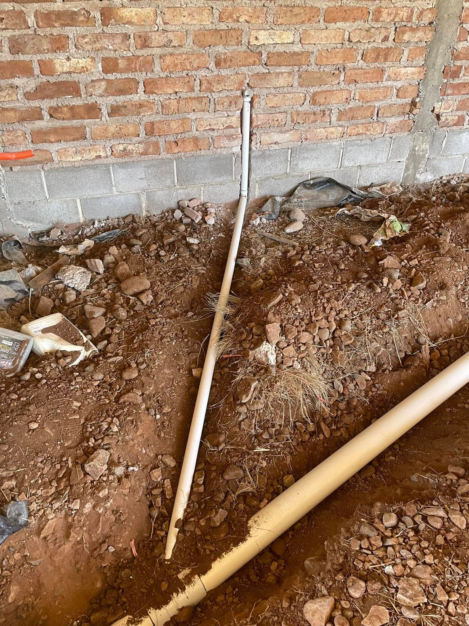 Excavation trench beside a brick wall with exposed pipes and muddy soil
