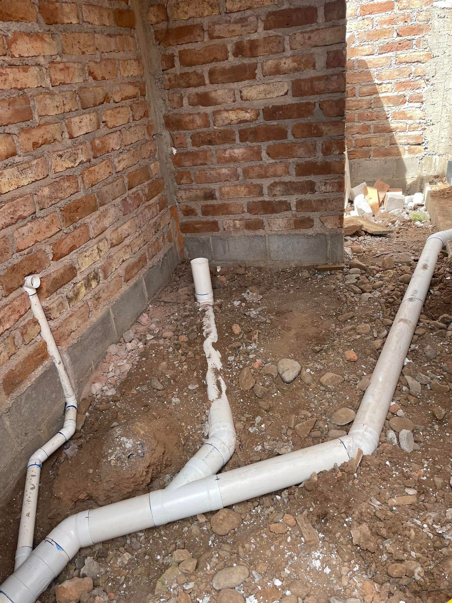 Exposed white PVC plumbing pipes along a brick wall in a dirt-floor crawlspace