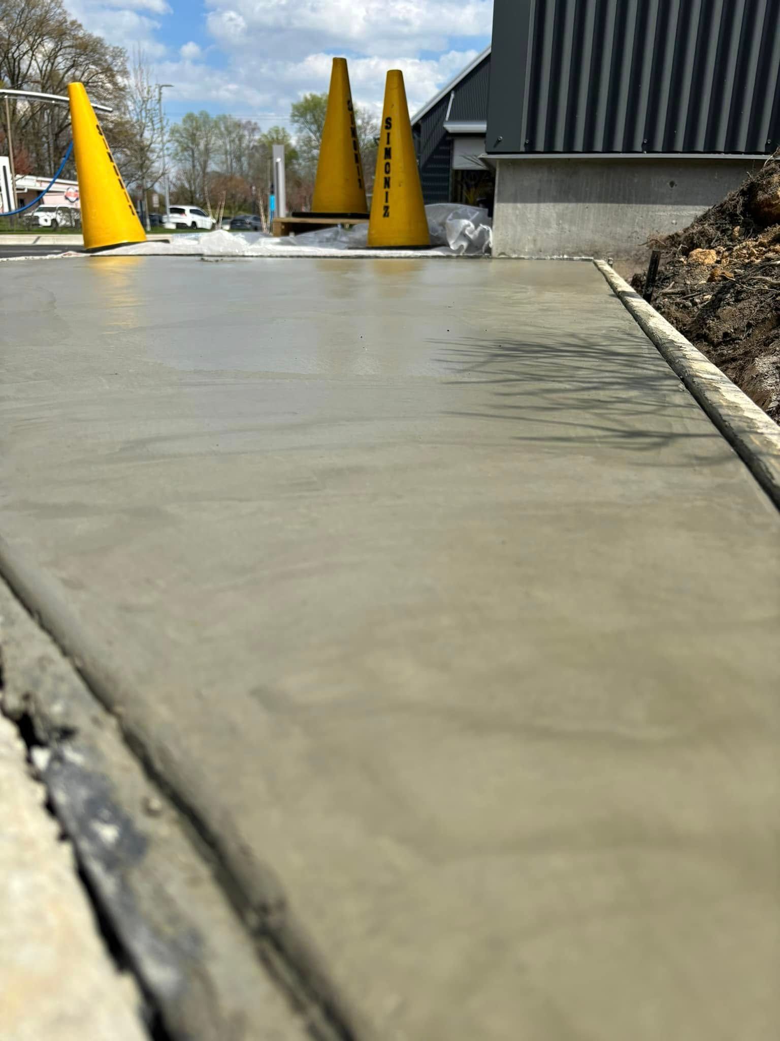 Wet concrete driveway with yellow traffic cones beside a building and trees.