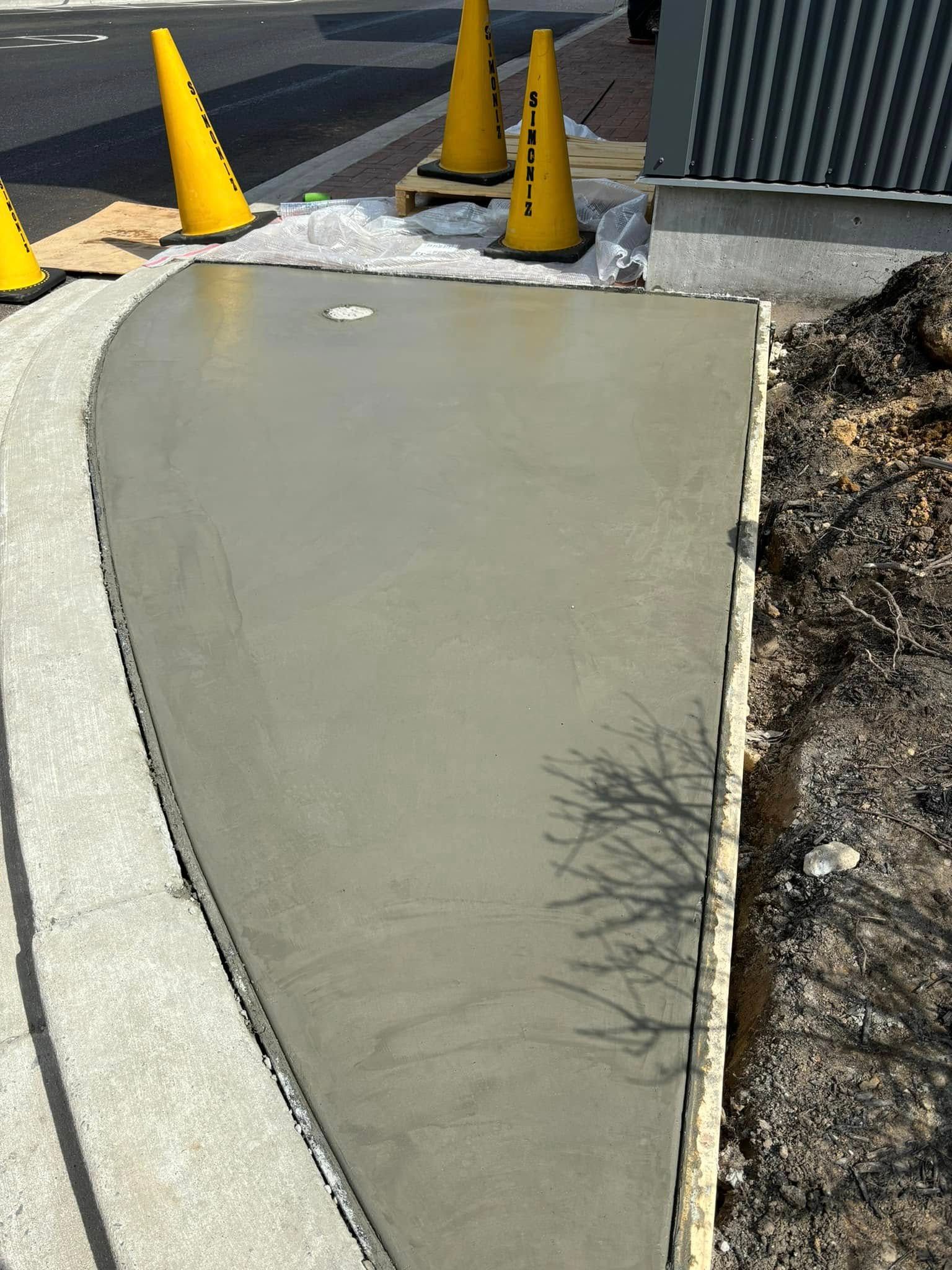 Concrete sidewalk ramp beside a curb and yellow traffic cones, with a dirt strip on the right.