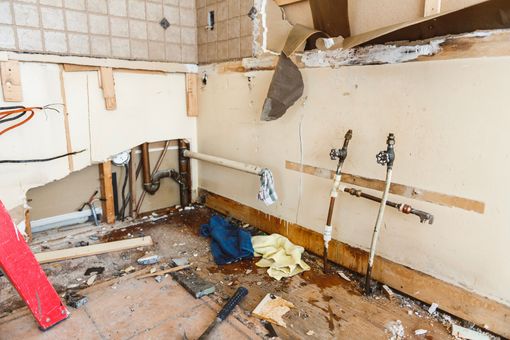 Damaged kitchen with broken cabinets, exposed pipes, debris, and peeling wall panels