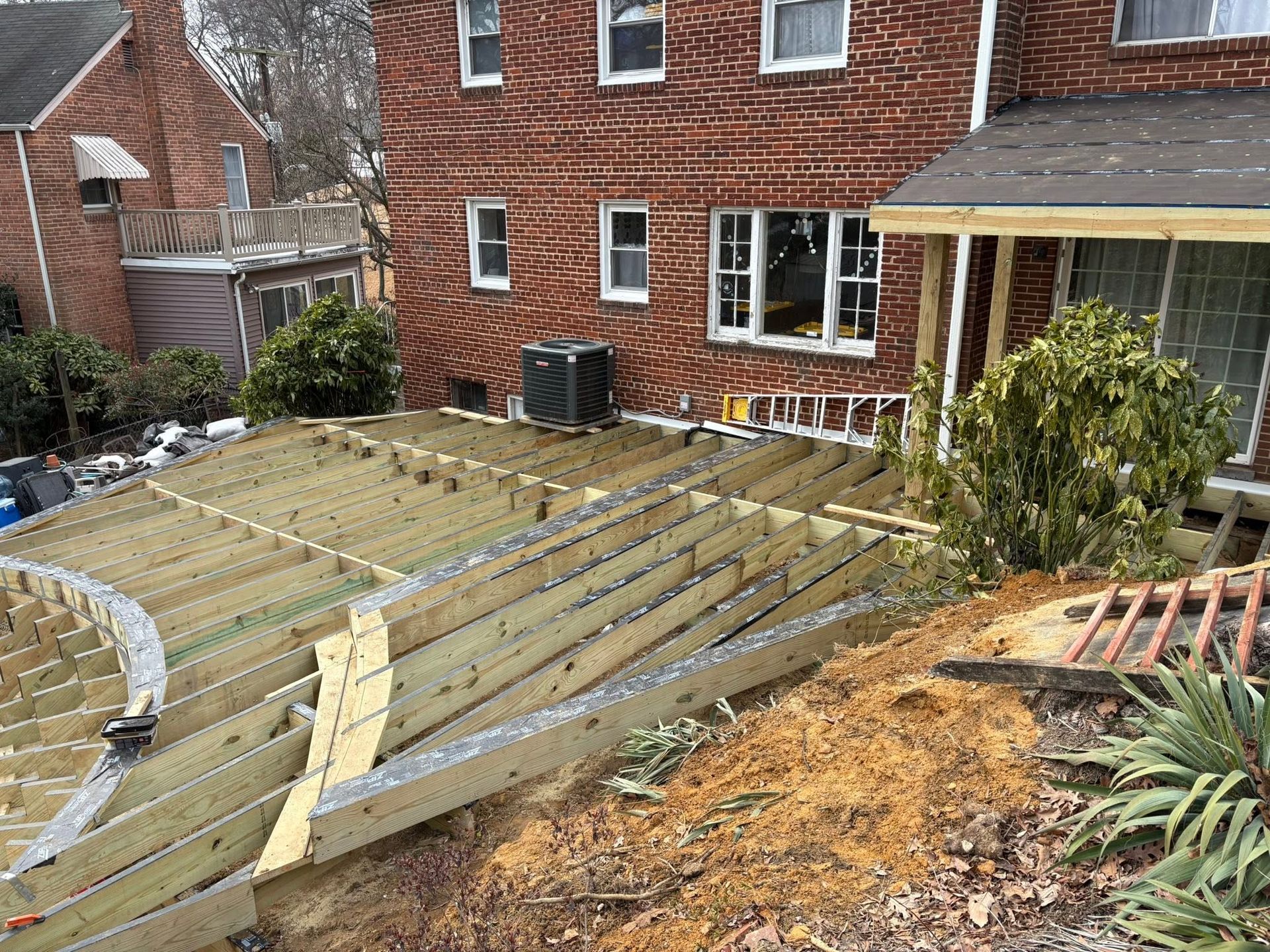 Backyard under construction with wooden deck framing beside a brick house and shrubs
