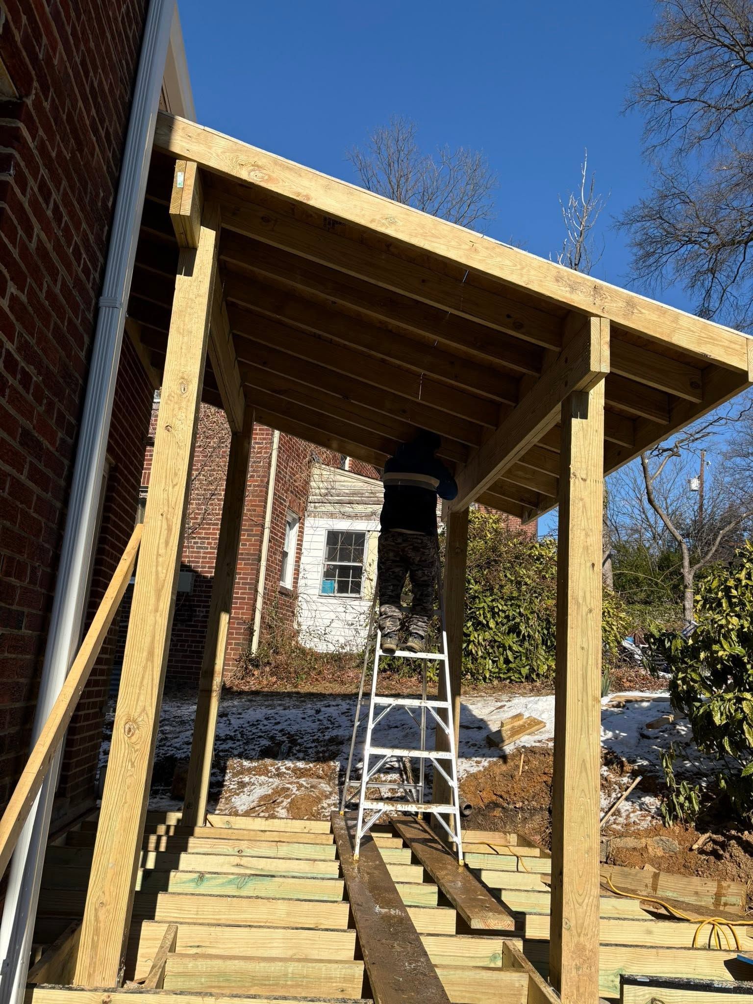 Wooden porch roof under construction beside a brick house, with a ladder and stacked lumber.