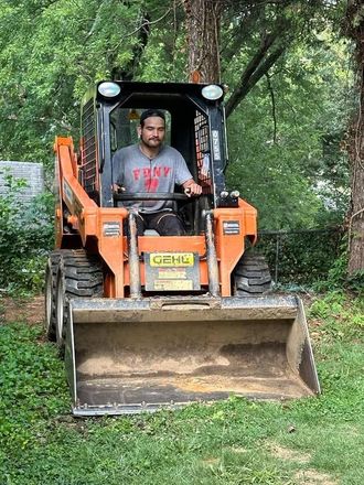 Person in an orange skid-steer loader clearing brush on a wooded path.
