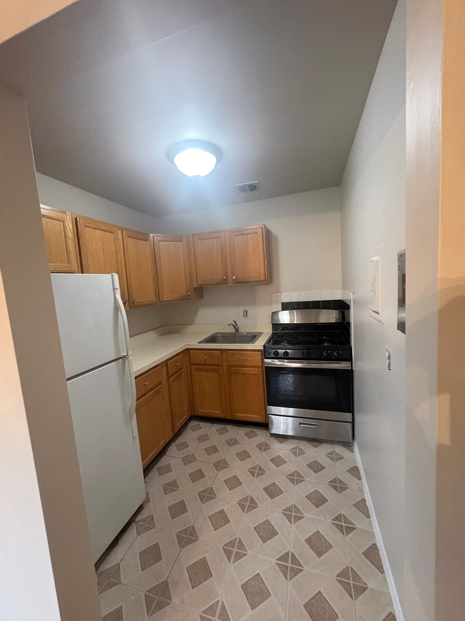 Small kitchen with wood cabinets, white fridge, black stove, and patterned floor under a ceiling light.