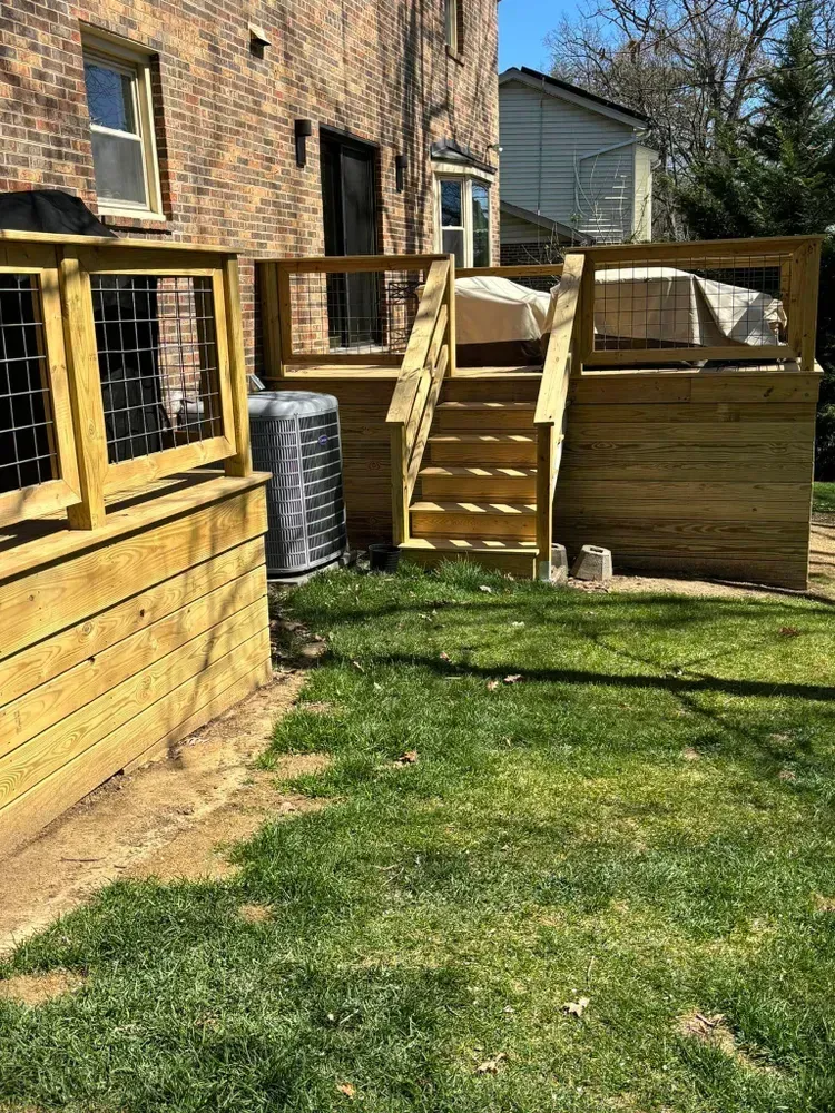 Wooden backyard deck with stairs, railings, and a window beside a brick house on a sunny lawn