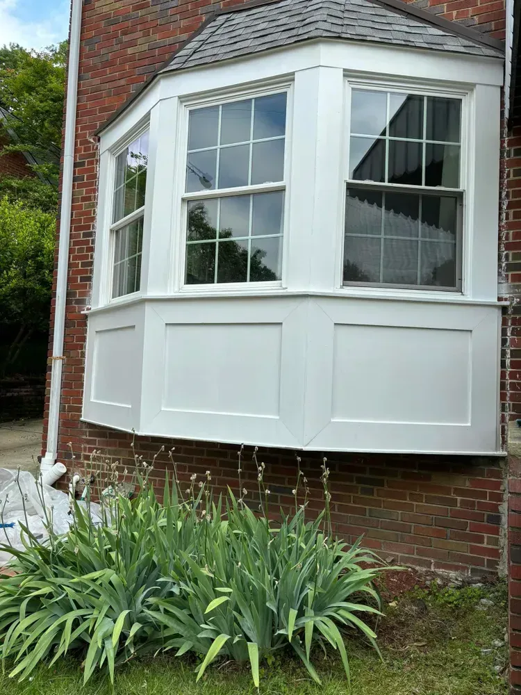 White bay window on a red brick house above green plants.