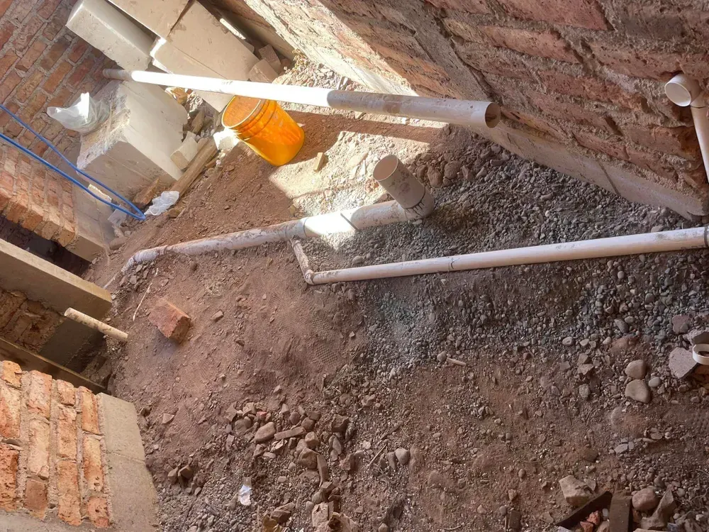 Narrow dirt trench with exposed pipes, rubble, and brick walls in a construction area