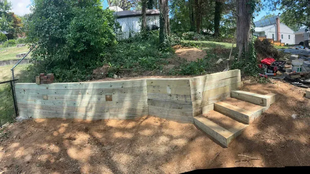 Concrete retaining wall with steps in a wooded yard, with houses and construction materials in the background