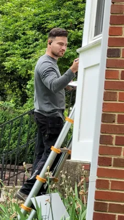 Man using a ladder to inspect or paint the outside of a white house beside a brick wall
