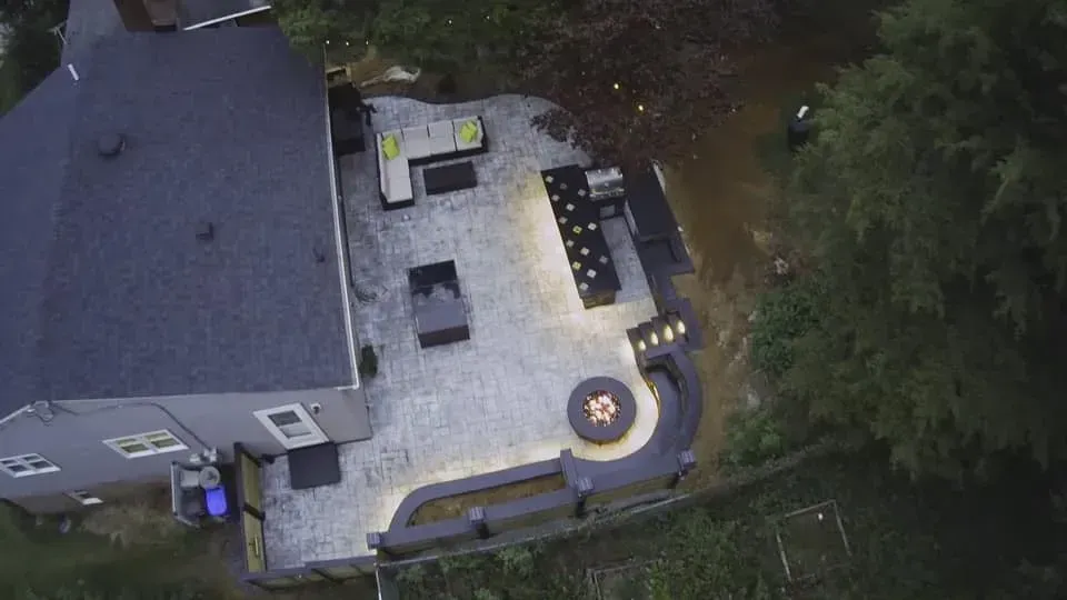 Aerial view of a house with a paved patio, outdoor dining area, fire pit, and surrounding trees