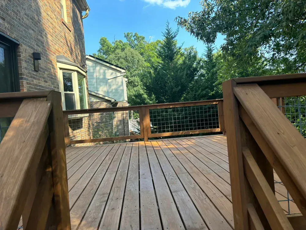 Wooden deck patio beside a brick house, with railing and trees in the background.
