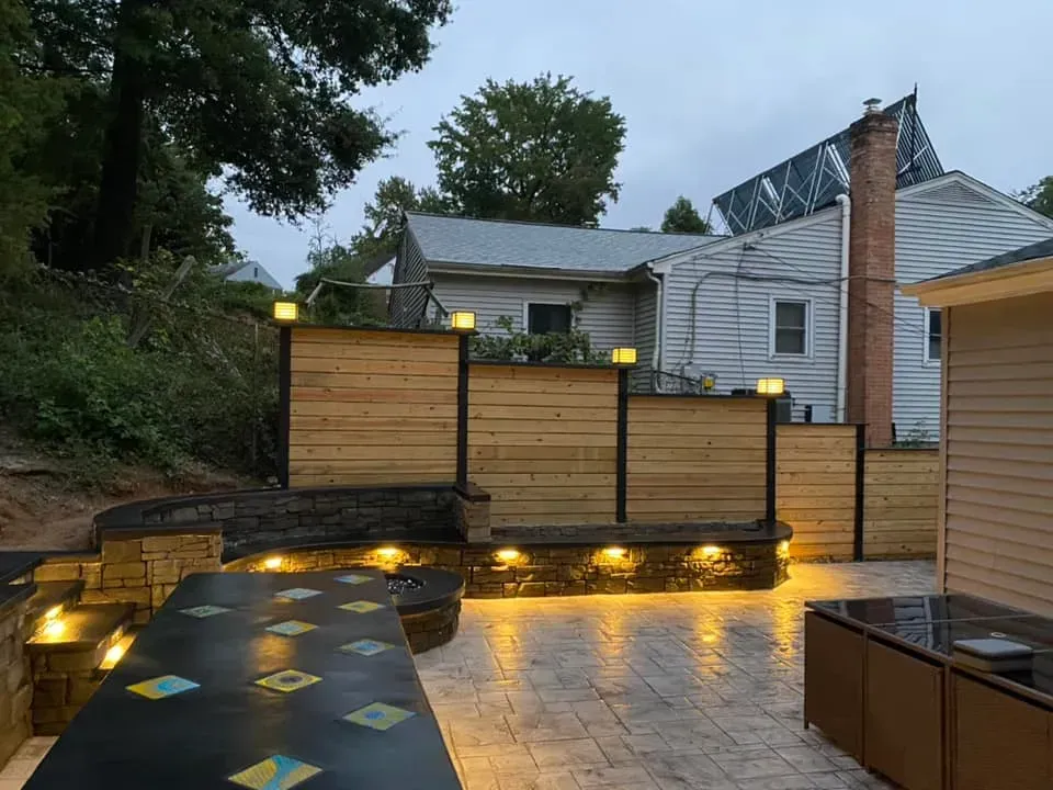 Lit backyard patio with wooden privacy screens, stone pavers, and warm step lights beside a house and chimney