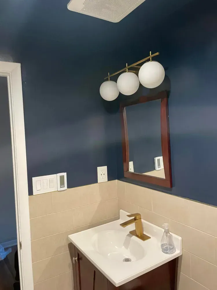 Small bathroom vanity with white sink, gold faucet, dark blue walls, and a round-bulb light fixture above mirror.