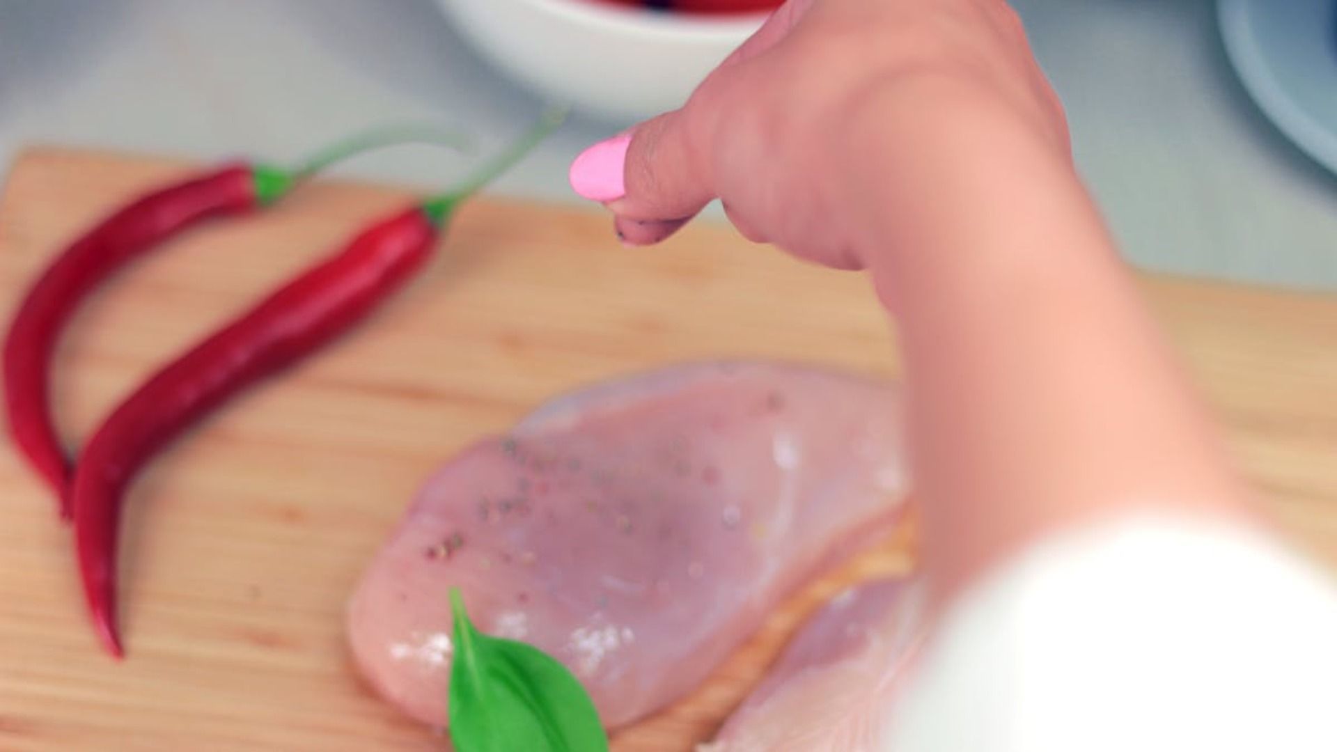Sazonando a mano pechugas de pollo crudas sobre una tabla de cortar, con chiles rojos al lado.
