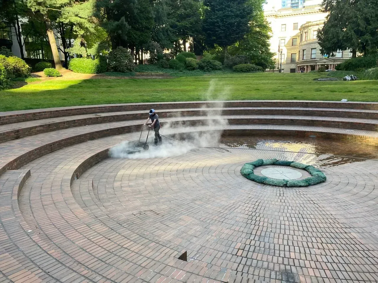 A person cleaning a circular brick fountain in a park, emitting steam. Terraced seating surrounds.
