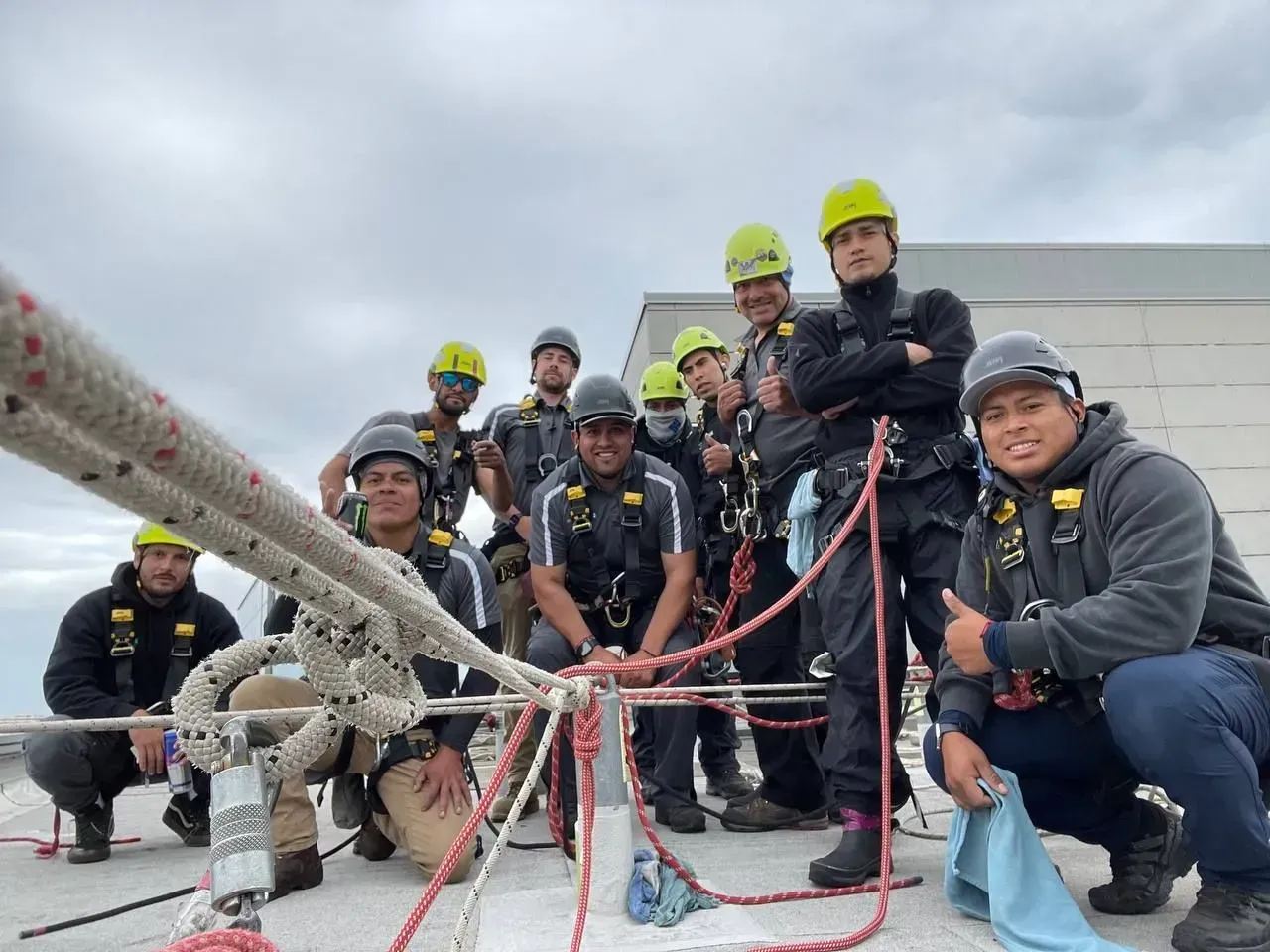 Group of workers on a rooftop with ropes, wearing helmets and safety harnesses, posing for the camera.