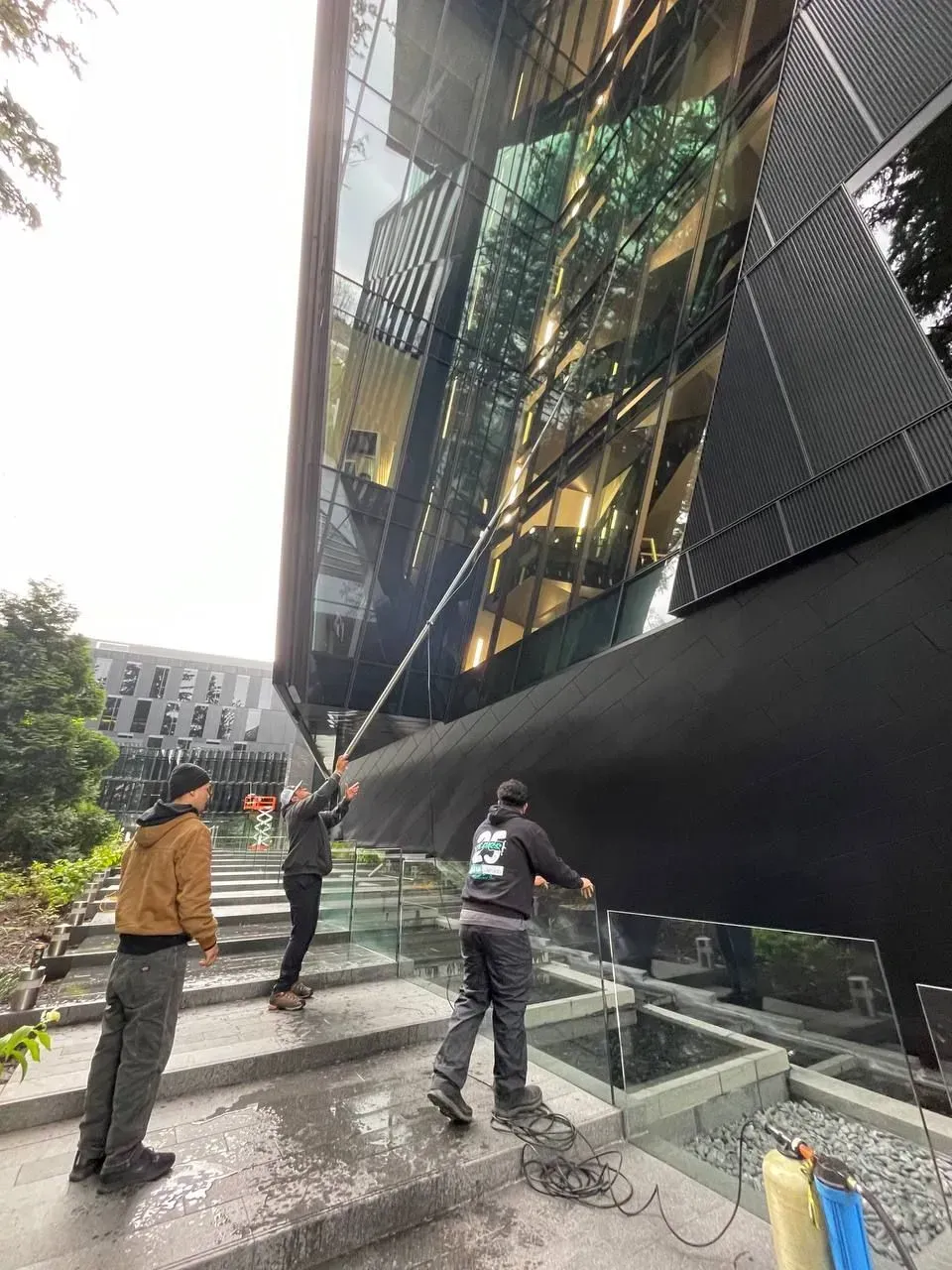 Three people cleaning the exterior of a modern building with reflective glass. One uses a long-handled brush.