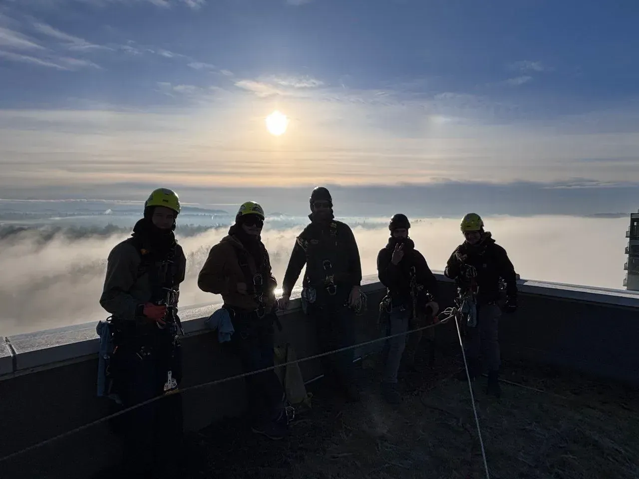 Five workers in safety gear on a rooftop, silhouetted against a sunrise over clouds.