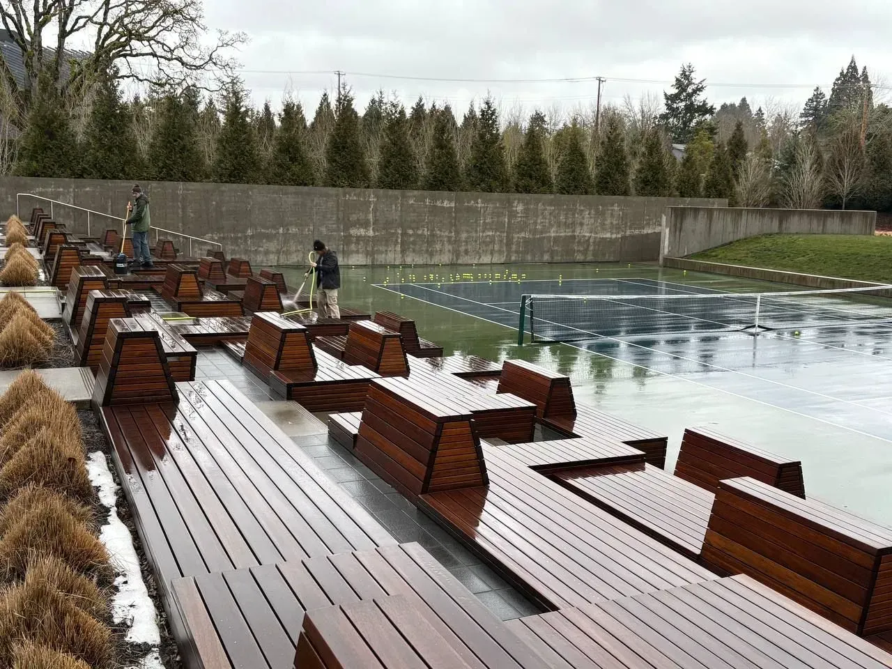 Outdoor seating area with wooden benches, tennis court, and two people. Cloudy day.