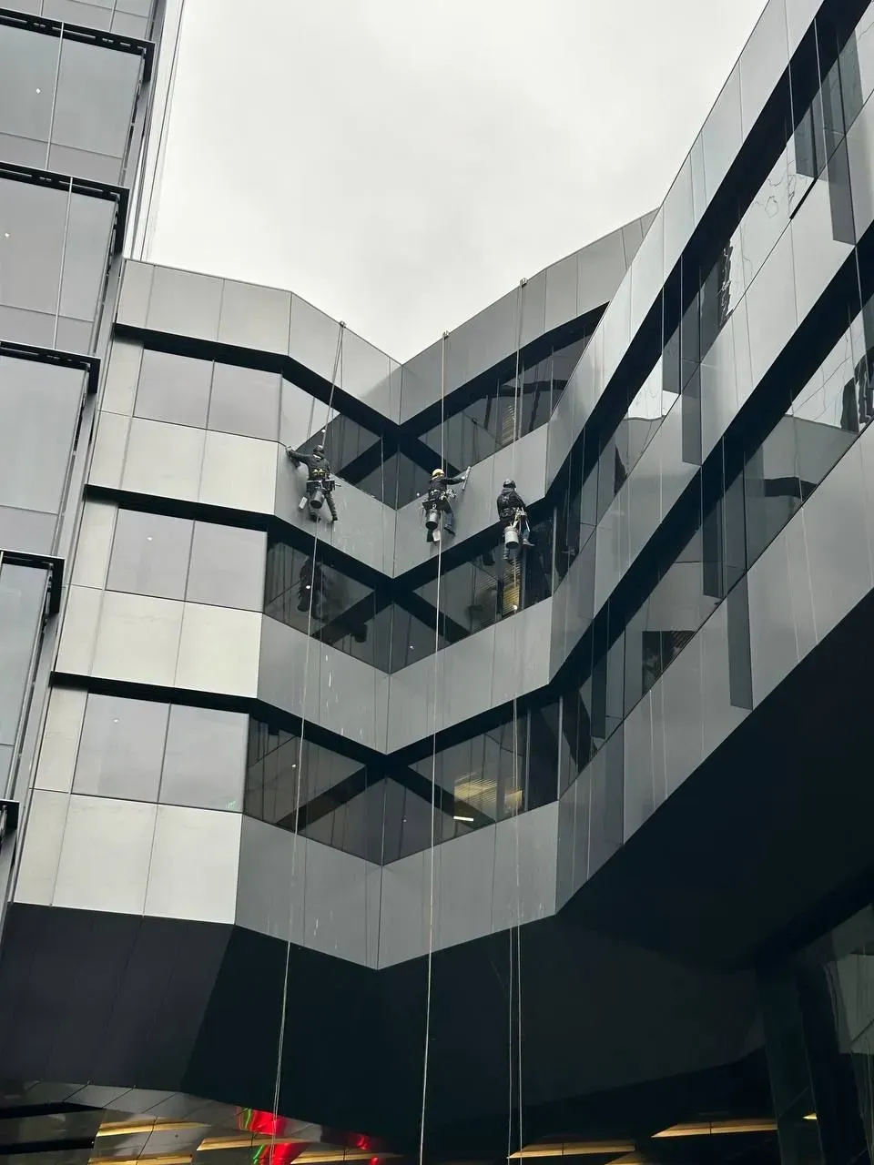 Window washers on ropes cleaning the exterior of a modern building with gray and black panels and reflective windows.