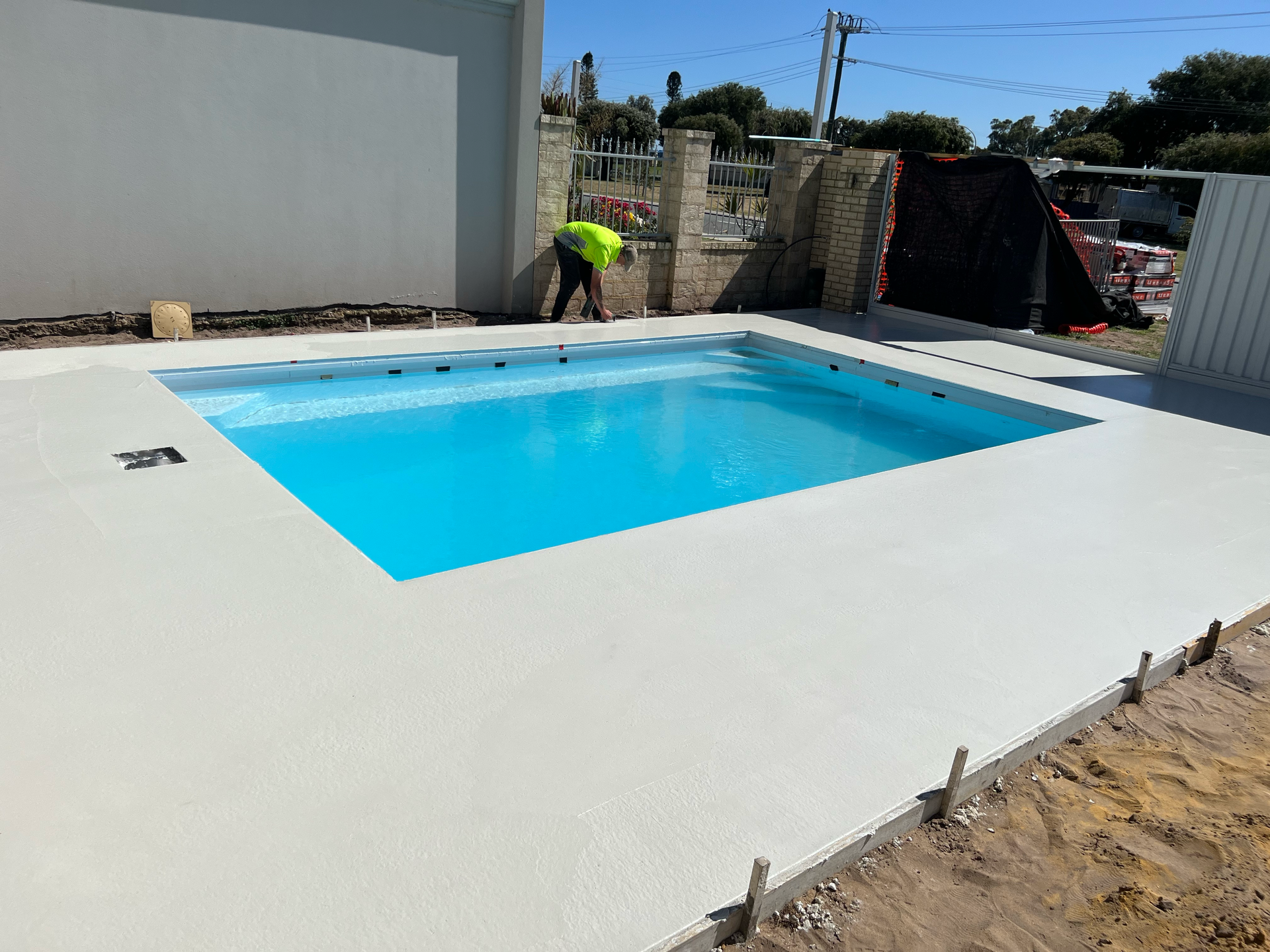 A backyard swimming pool with a bright blue water, white deck, and a worker in a neon vest nearby.