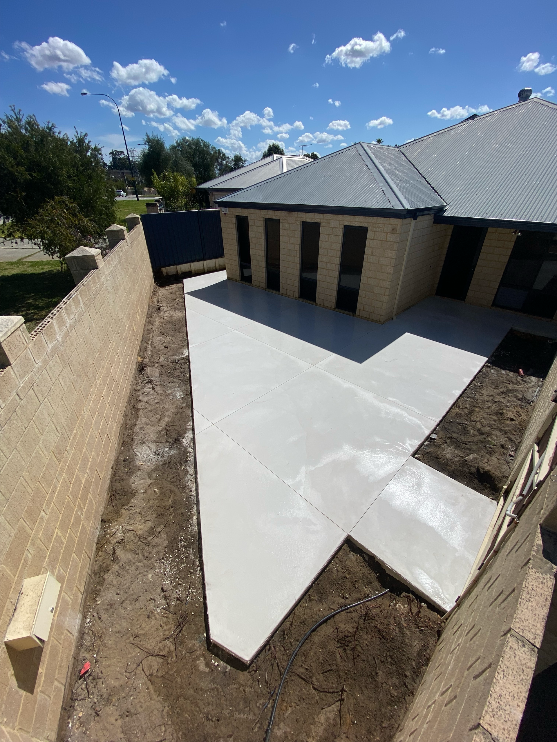Backyard patio concrete slab beside a tan house and block wall under a blue sky