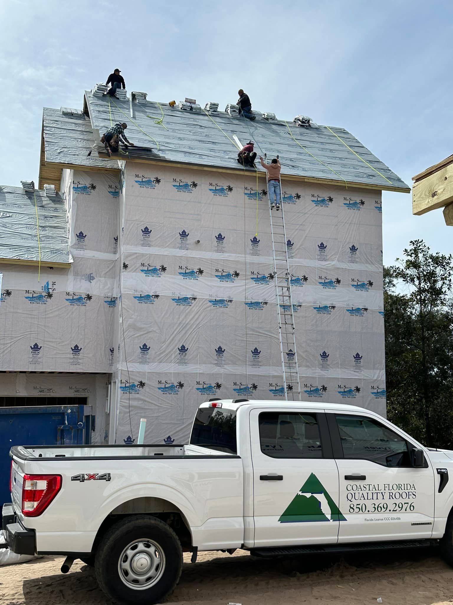 White work truck parked beside a house wrapped in insulation and roofing under construction