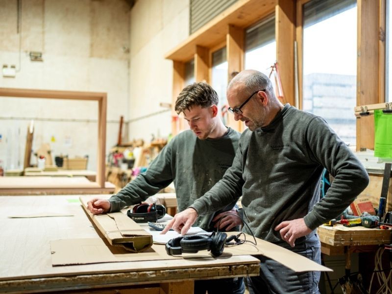Two people measuring wood at a workbench in a workshop