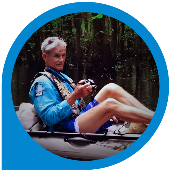 Man in kayak taking photo in a swamp. He wears blue shirt, life vest, shorts; trees behind.