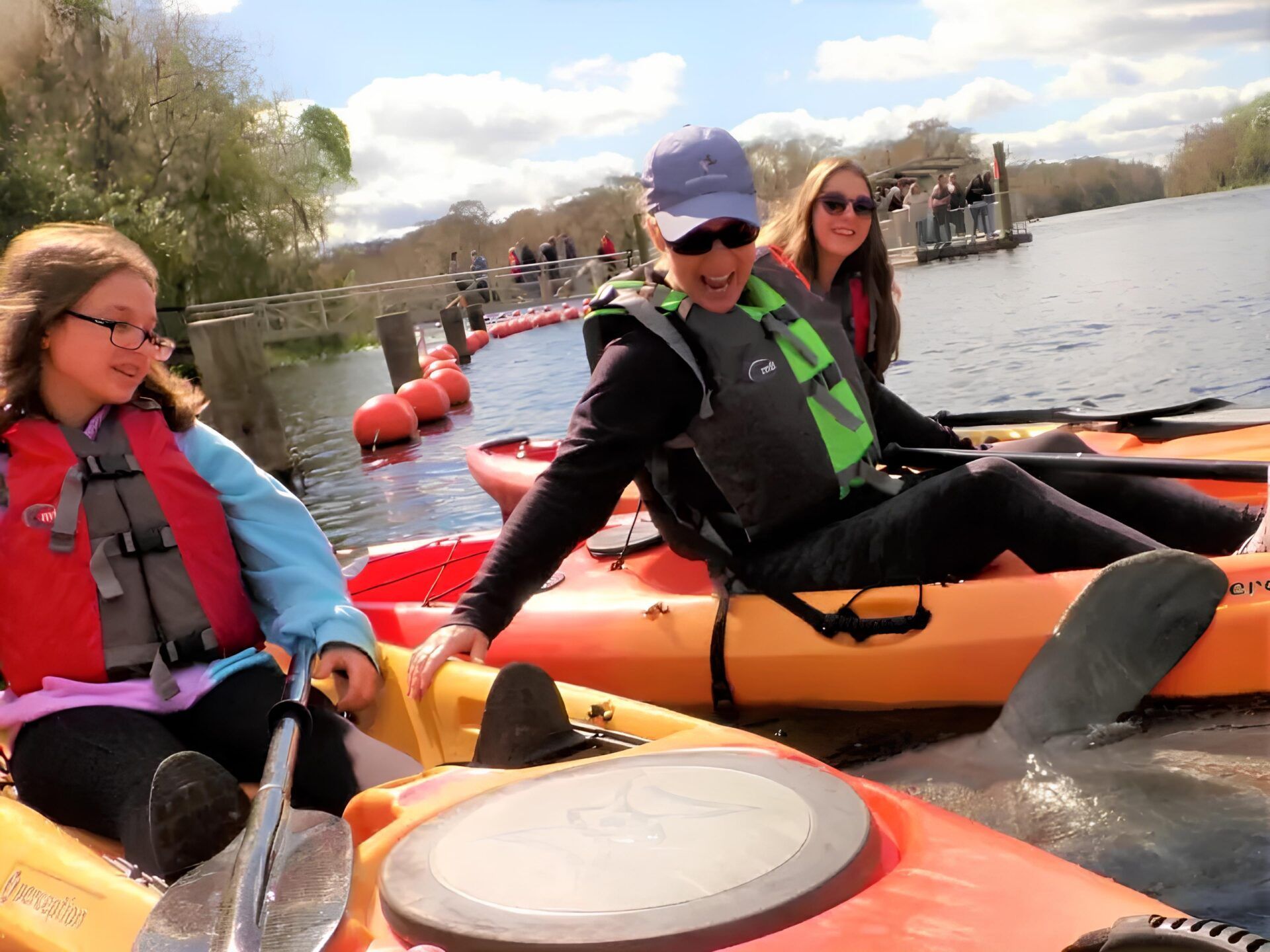Three people in kayaks on water near a dock, wearing life vests. Sunny day.