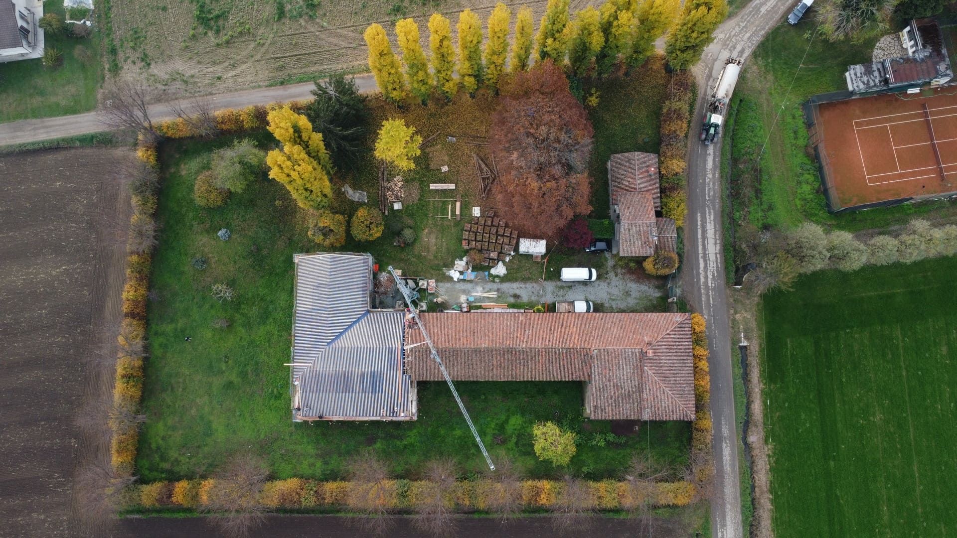 Vista aerea di una proprietà rurale con edifici, un campo da tennis e campi circostanti; prevalentemente verdi e marroni.