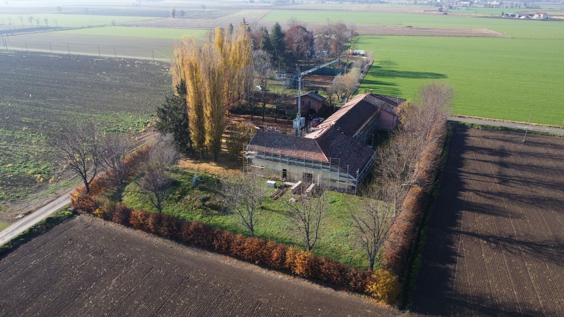 Vista aerea di un edificio agricolo con tetto di tegole rosse, circondato da campi e da una siepe.