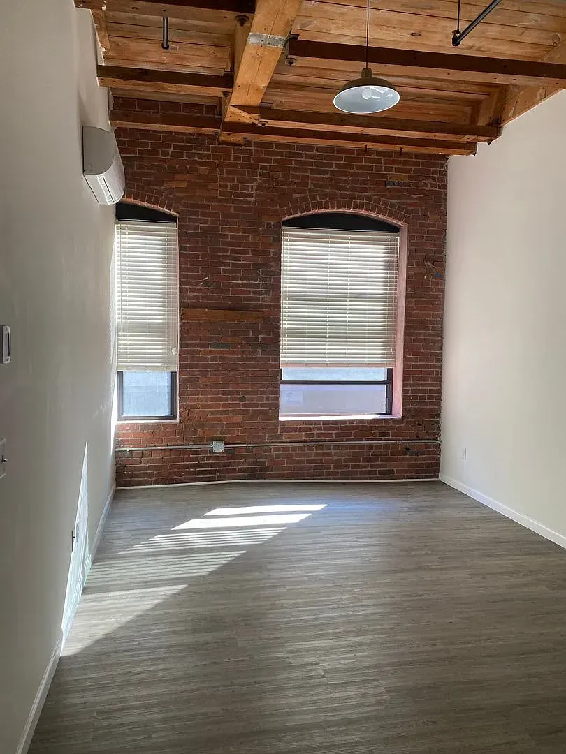 Empty room with exposed brick wall, windows with blinds, wood beams, and gray flooring.