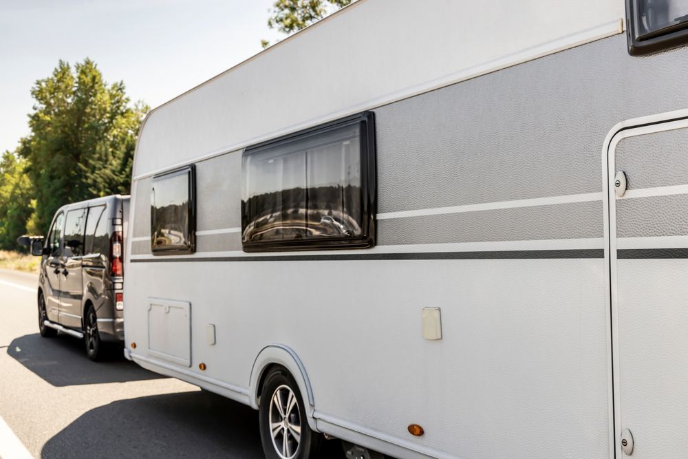 White RV trailer hitched to a van parked on a sunlit street beside trees