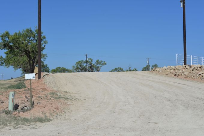 Dirt road rising over a hill under a clear blue sky, with sparse trees and utility poles