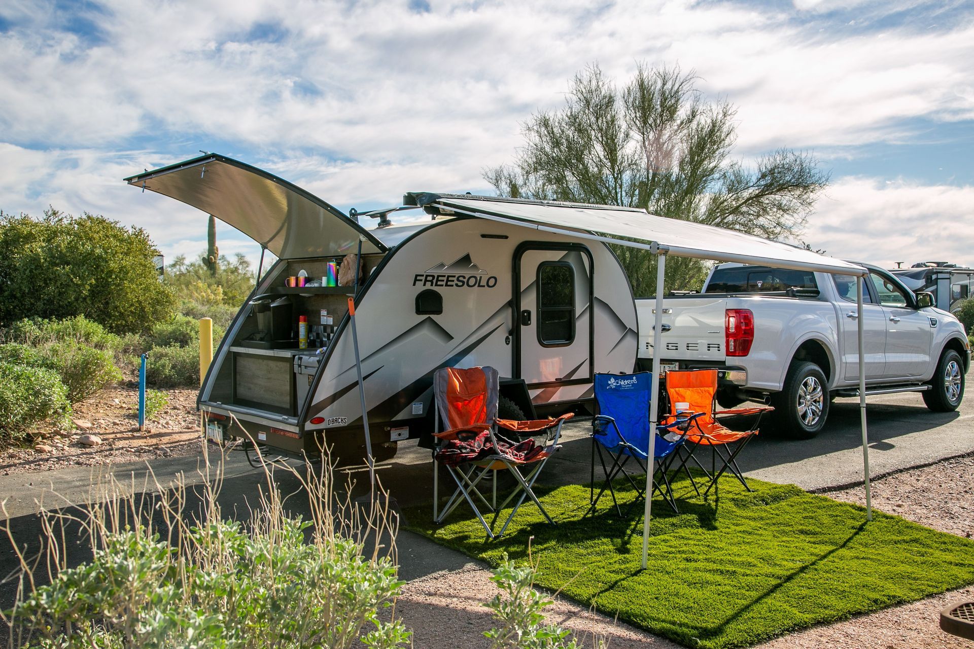 White camper trailer parked at a campsite with awning, chairs, and a silver pickup truck beside it.