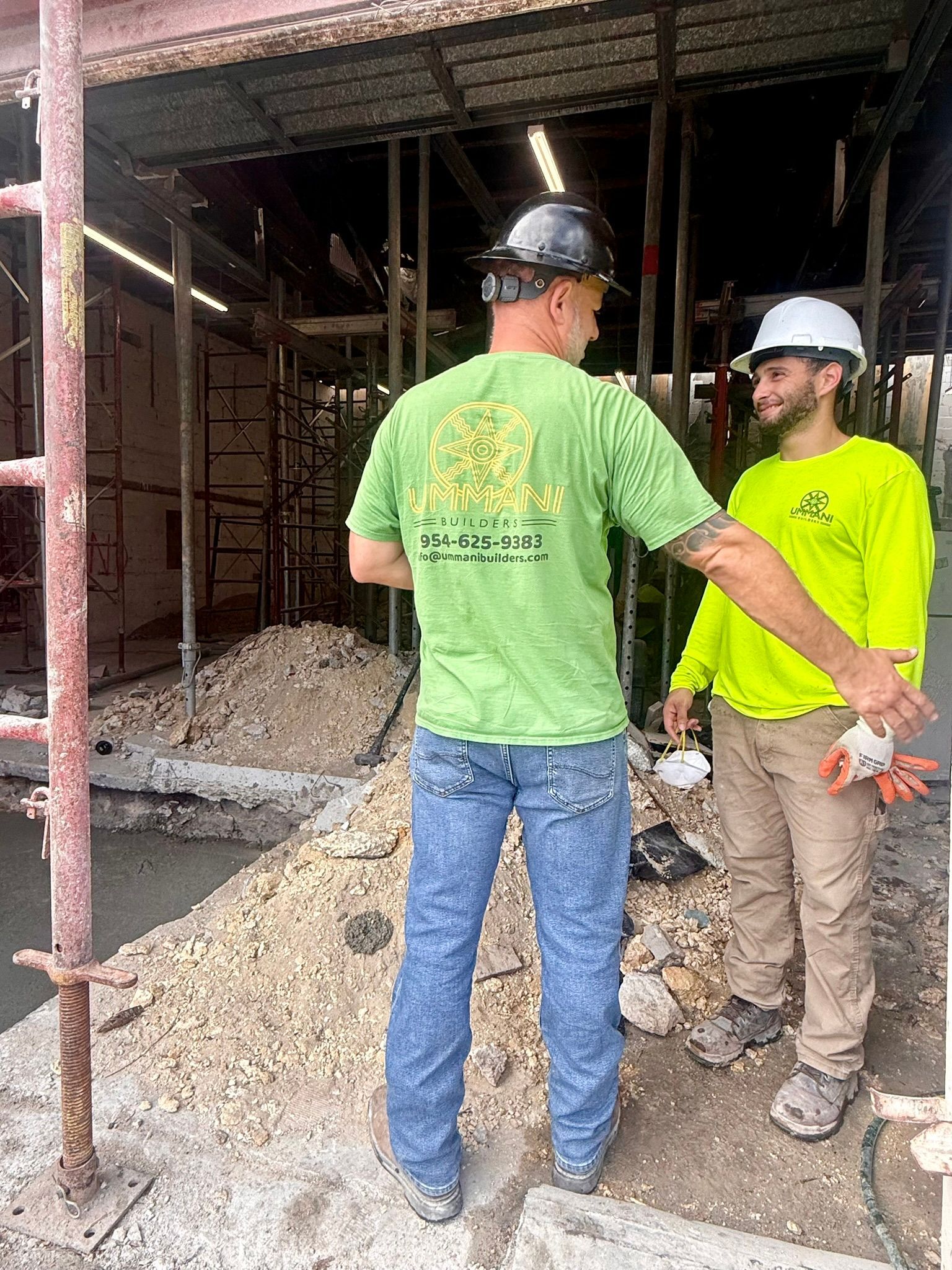 Two workers in safety gear talk at a construction site with scaffolding and gravel.