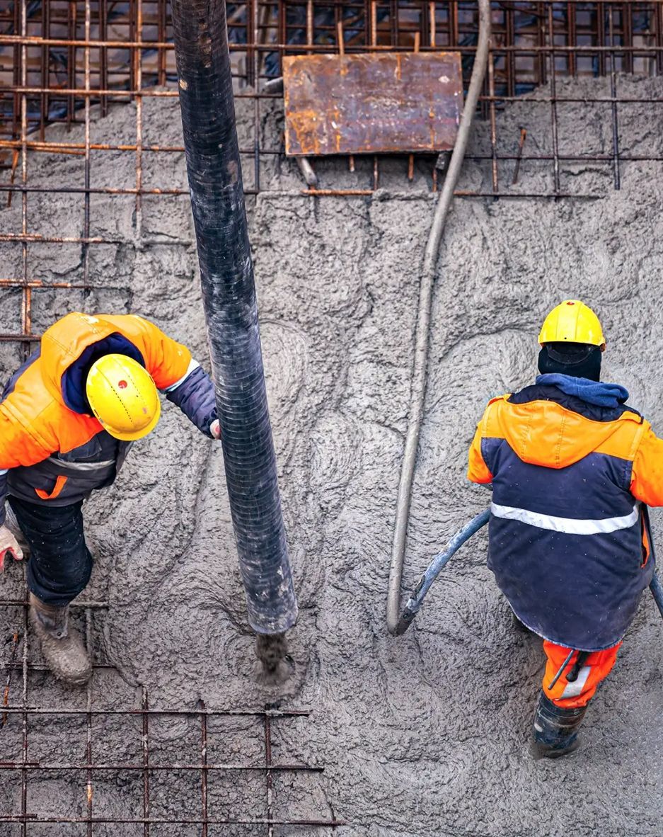 Two construction workers in high-visibility gear work on a site, pouring concrete into a metal-reinforced foundation.