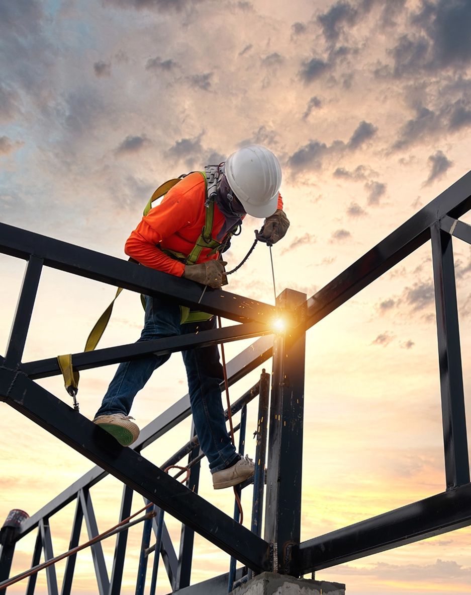 A construction worker in a hard hat and safety harness welds a metal frame against a sunset sky.