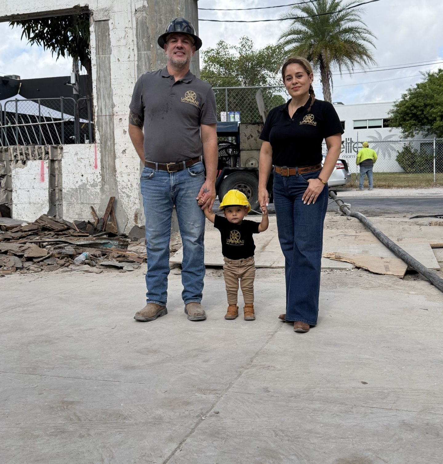 A family of three stands on a concrete construction site, wearing matching logo shirts, boots, and safety gear.