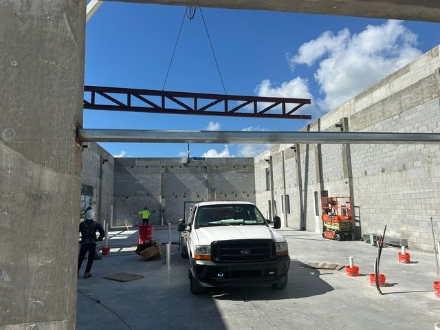 A construction site with a truck parked on a concrete floor as a metal truss is lifted by a crane overhead.