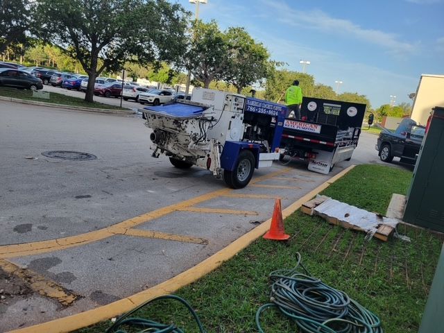 A white industrial machine hitched to a black trailer in a parking lot, with a traffic cone and hoses on the grass.