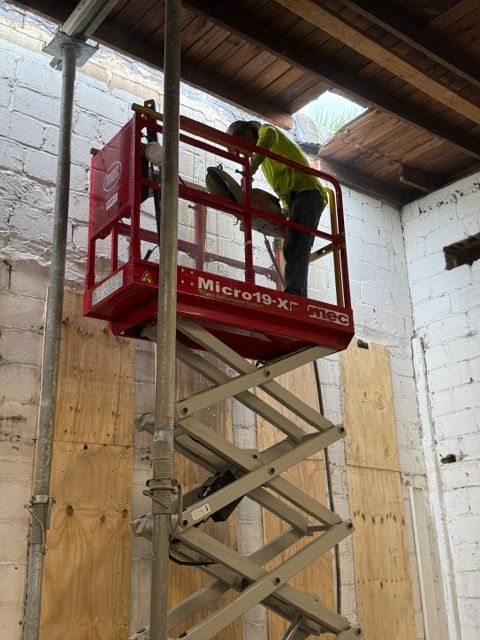 A worker in a bright yellow shirt stands on a red scissor lift, working on the ceiling of an indoor construction site.