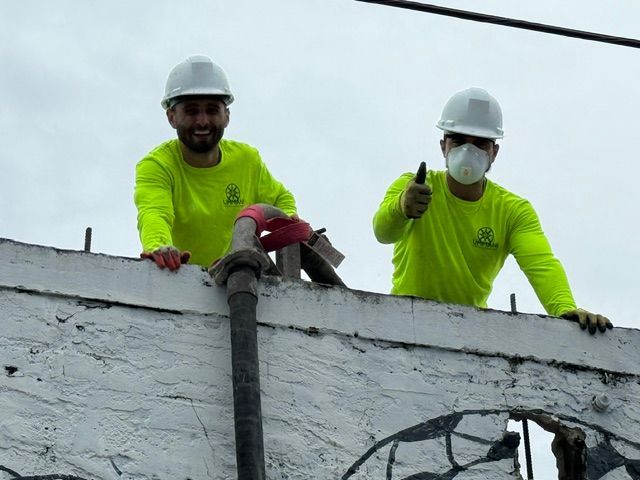 Two workers in hard hats and neon shirts operate a concrete hose from the top of a white, textured wall.