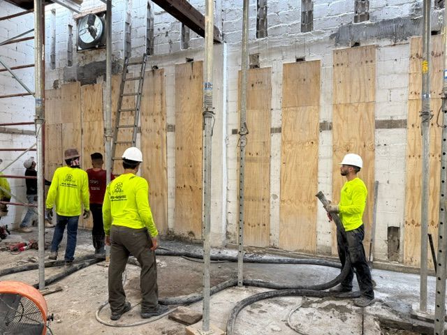 Workers in yellow safety shirts use a hose to fill plywood-covered wall sections in a construction site.